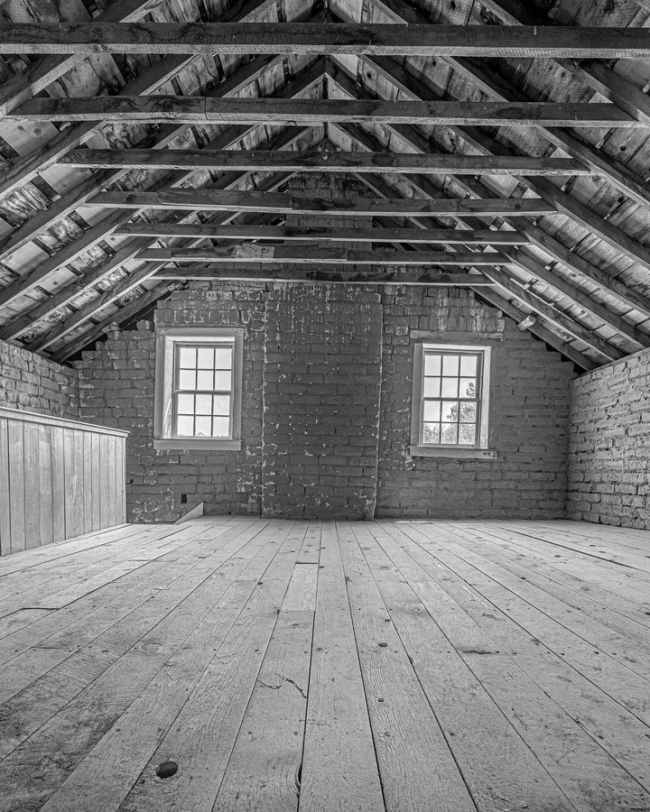 Leerer Dachboden mit sichtbarem Dachstuhl und Kehlbalken über Holzdielen, hinten ein Backsteingiebel mit zwei Fenstern